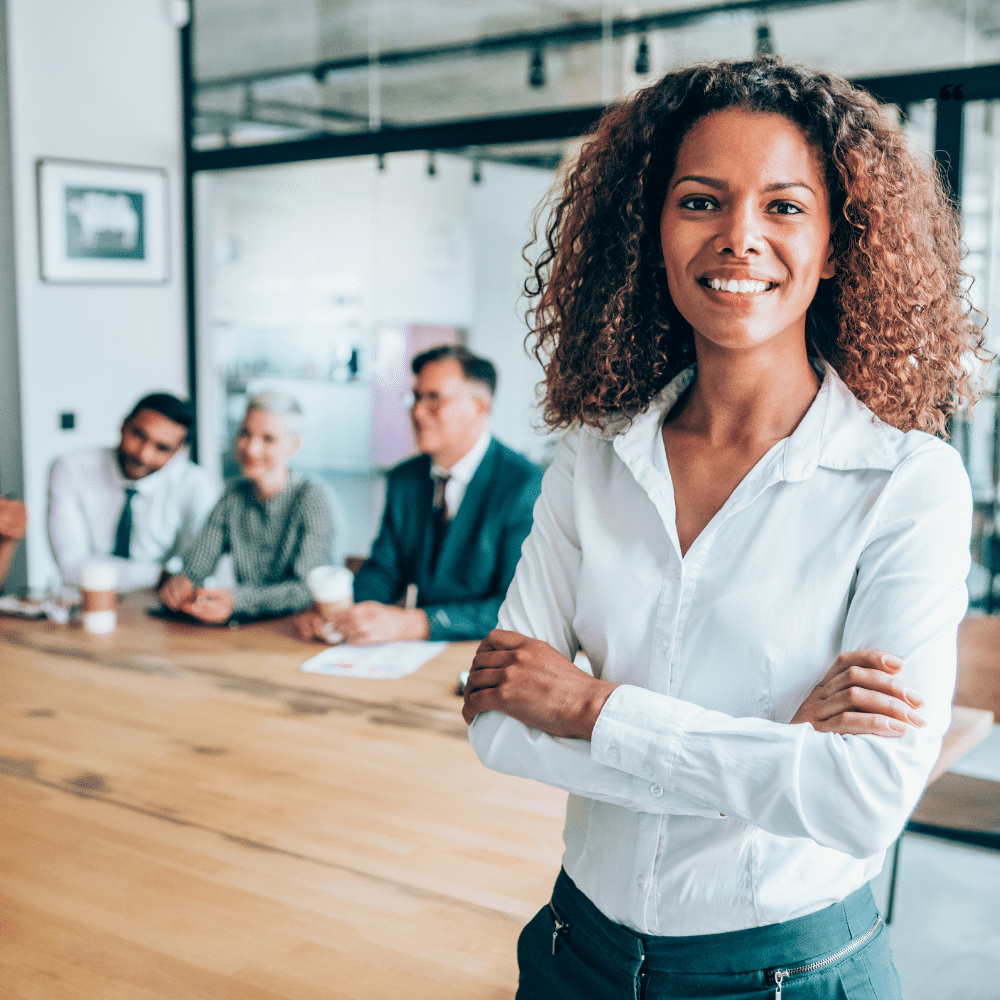 A confident businesswoman with curly hair, wearing a white blouse, stands with her arms crossed in the foreground while a diverse group of professionals sits at a conference table in the background.